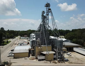Industrial grain processing facility with tall metal silos, conveyor towers, storage buildings, trucks, and a rural road beneath a partly cloudy sky.