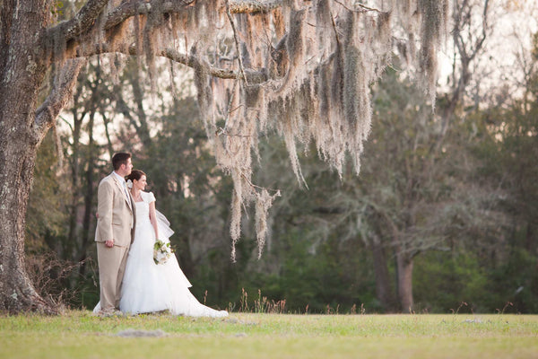 Bride and groom stand together beneath a moss covered oak tree in a grassy landscape with the bride holding a small bouquet