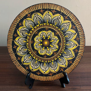 Intricately painted circular decorative plate with a black, gold, and yellow floral mandala pattern, displayed upright on a small stand against a wall.