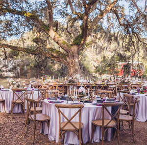 Outdoor wedding reception with round tables, lavender linens, wooden crossback chairs, floral centerpieces, and candles beneath a sprawling oak tree.