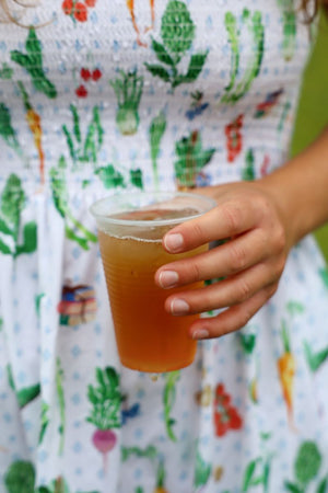 Person wearing a colorful botanical apron holds a clear cup of amber iced tea outdoors”】【