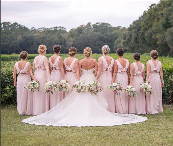 Bride and bridesmaids pose in a lush green field, viewed from behind, with pastel bouquets and a flowing white wedding dress train.