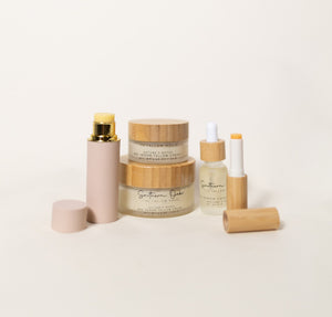 Assorted beige skincare containers with wooden lids, including stacked jars, a pump bottle, dropper bottle, and open cylindrical packaging arranged on a light background