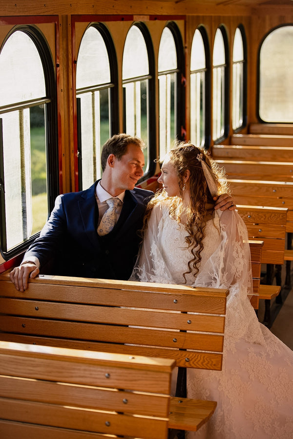 A bride and groom in wedding attire sit together on a wooden passenger trolley, smiling beside tall arched windows and sunlit countryside.
