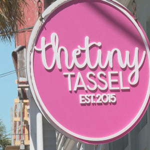 Round pink hanging sign with white lettering that reads The Tiny Tassel and Est. 2015, displayed outside a colorful storefront.
