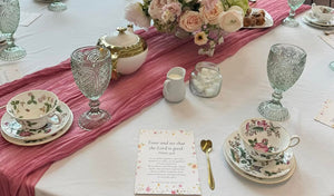 Elegant tea table setting with floral china, crystal goblets, gold flatware, a rose centerpiece, tea service, and a printed card on a pink runner