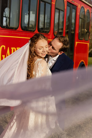 Bride and groom share a kiss beside a bright red trolley, with the bride's flowing veil and wedding dress visible in the foreground.