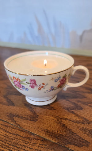 Floral porcelain teacup holding a small lit candle on a wooden table, with a softly blurred blue and beige background.