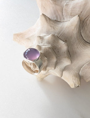 Silver ring with a smooth oval purple stone resting on the ridges of a large textured seashell against a light background