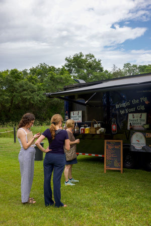 Three women stand in line outside a food truck in a grassy area with trees in the background under a partly cloudy sky.