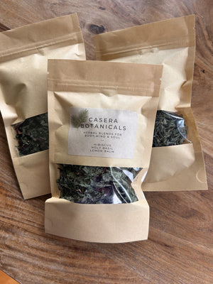 Three kraft paper pouches of loose herbal blends arranged on a wooden table, with the center label reading Casera Botanicals and listing hibiscus, holy basil, and lemon balm.