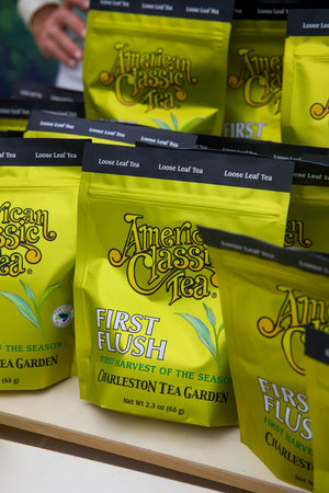 Bright gold American Classic Tea pouches labeled First Flush and Charleston Tea Garden displayed on a shelf with loose leaf tea labels and green tea leaves