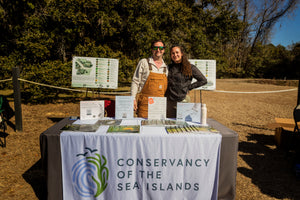 Two women stand behind a Conservancy of the Sea Islands information table outdoors, with brochures, signs, and nature materials displayed in a sunny park setting.