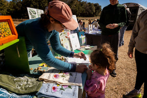At Charleston Tea Garden, a woman helps a young girl look at printed T shirts on an outdoor table beside colorful displays and visitors.