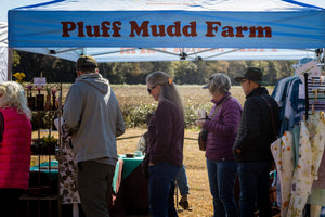 Visitors browse handmade goods and clothing at the Pluff Mudd Farm market booth, with a sunlit field visible behind the canopy.