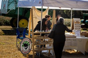 Visitors browse jars of tea and packaged assortments at a Charleston Tea Garden outdoor market stall beneath a canopy.