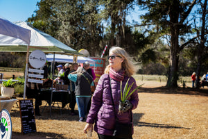 Woman in a purple jacket and scarf carries a small potted plant at an outdoor garden event with vendor tents, signs, and people beneath large trees.