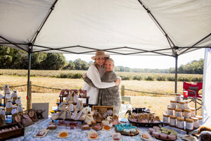 Two women smile behind an outdoor tea stand displaying jars, packaged tea assortments, soaps, and baked goods beneath a canopy beside open fields.