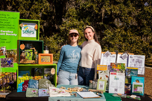 Two women stand beside an outdoor table displaying brochures books artwork and colorful handmade items at a community information booth surrounded by trees