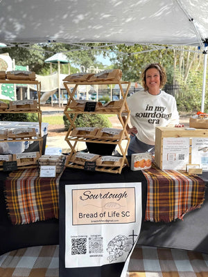 Woman standing at an outdoor market stall displaying sourdough breads on wooden shelves, with a Bread of Life SC sign, plaid tablecloth, and canopy overhead