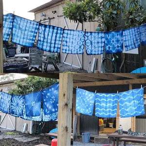 Several blue and white tie-dye fabric squares hanging on clotheslines outdoors near wooden structures and garden tools.
