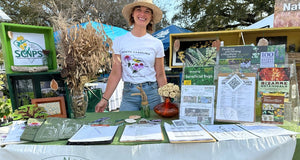 Woman in a straw hat and South Carolina T shirt stands behind an outdoor information table with brochures, plants, and colorful community displays