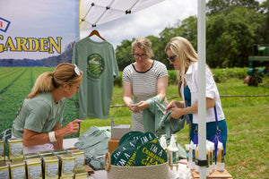 Visitors browse Charleston Tea Garden apparel, hats, tea tins, and gift items at an outdoor market booth while staff assist them.