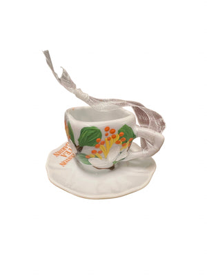 White ceramic teacup ornament with colorful floral accents on a matching saucer and a silver hanging ribbon for holiday display
