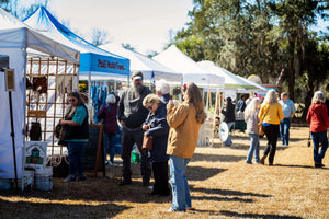 People exploring and shopping at outdoor market stalls on a sunny day with tents showcasing various items and trees in the background.