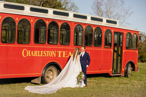 Bride and groom pose beside a bright red Charleston Tea tour bus in a grassy field, with the bride’s long veil spread across the ground