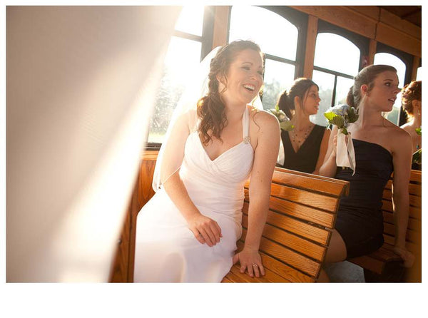 Bride in a white dress laughs while seated on a wooden bench beside bridesmaids inside a sunlit vehicle with large windows.