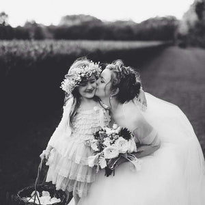 Black and white wedding portrait of a bride kissing a smiling flower girl outdoors, with the bride holding a bouquet and wearing a flowing veil.