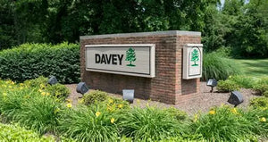 Brick entrance sign displaying the word Davey and a green tree logo, surrounded by flowering plants, ornamental grasses, shrubs, and mature trees