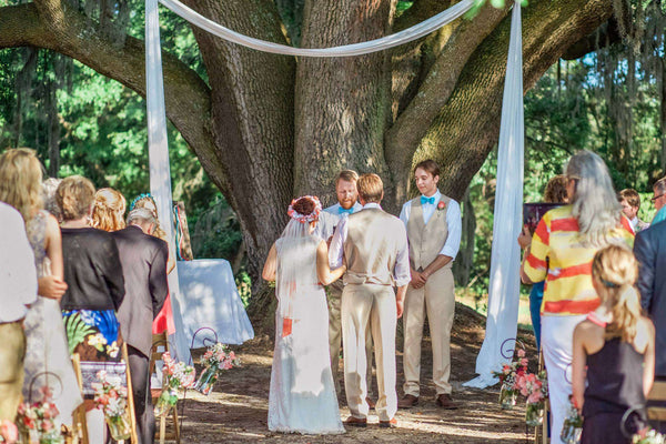 Outdoor wedding ceremony beneath a large tree, with the bride and groom standing before an officiant as guests gather around a flower decorated arch.