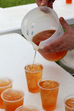 Hands pouring iced tea from a clear pitcher into a plastic cup filled with ice on a white table, with several other iced tea cups nearby outdoors.