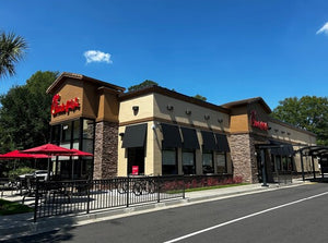 A modern restaurant building with red umbrellas, outdoor patio seating, stone walls, black awnings, and a clear blue sky.